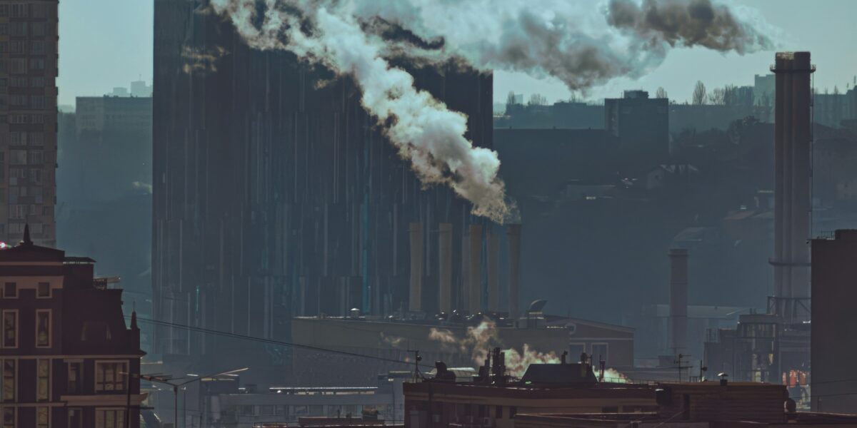 Industrial cityscape with multiple smokestacks releasing smoke into a hazy sky representing how GDP growth capitalism harms public health across food work environment housing and healthcare