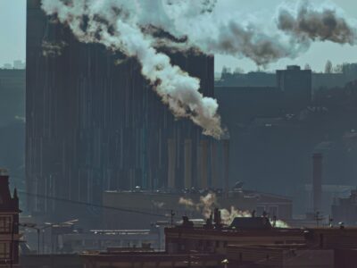 Industrial cityscape with multiple smokestacks releasing smoke into a hazy sky representing how GDP growth capitalism harms public health across food work environment housing and healthcare